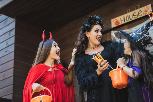 Animado mulher segurando doces perto de filhas no diabo e bruxa halloween trajes — Fotografia de Stock