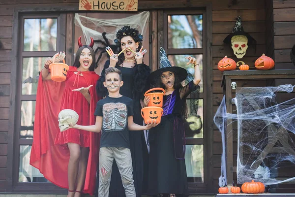 Crianças com mãe em trajes de Halloween em pé com baldes, doces e crânio no alpendre com decoração — Fotografia de Stock