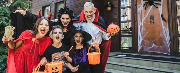Family in spooky costumes holding halloween attributes while growling at camera, banner — Stock Photo
