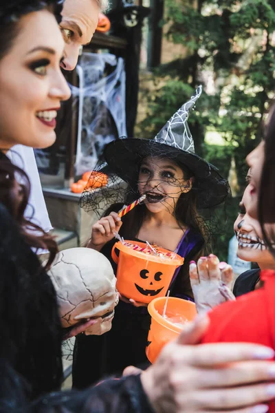 Счастливые дети в жутких костюмах на Хэллоуин едят конфеты рядом с размытыми родителями — стоковое фото
