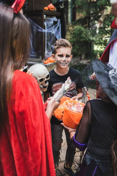 Garçon heureux en costume d'Halloween près de la famille floue avec des seaux de bonbons — Photo de stock