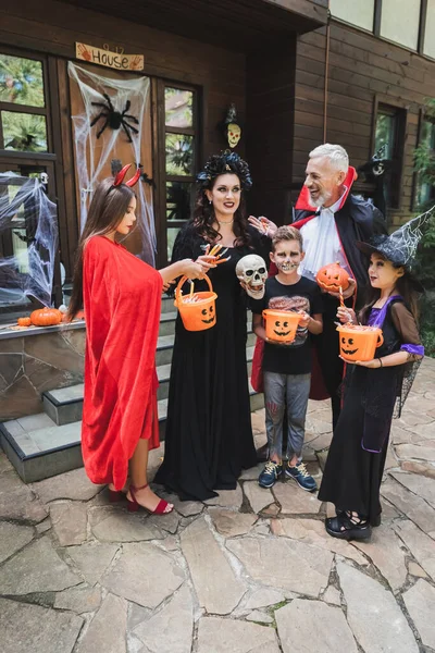 Família alegre em fantasias assustadoras segurando baldes de doces perto da casa de campo com decoração de Halloween — Fotografia de Stock
