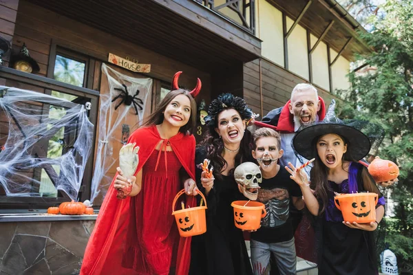 Cheerful family in spooky halloween costumes grimacing at camera near house — Stock Photo