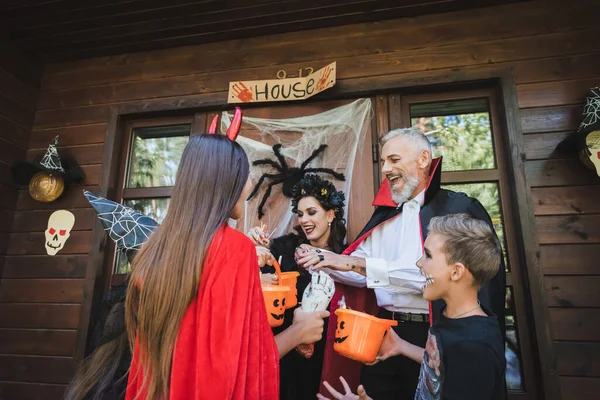 Crianças animadas em trajes de Halloween segurando baldes com doces perto de vizinhos no alpendre — Fotografia de Stock