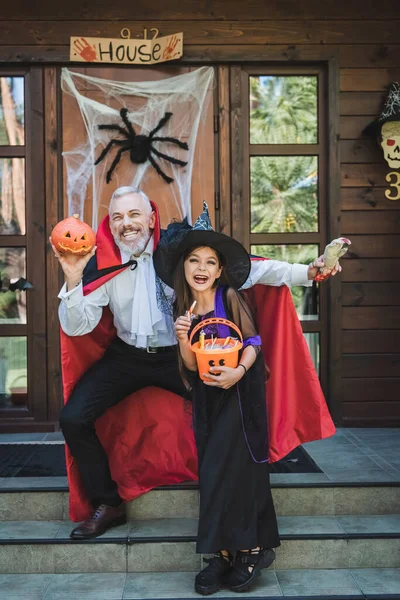 Cheerful girl and father in halloween costumes smiling on house porch with decoration — Stock Photo