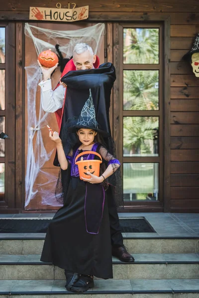 Maduro hombre e hija en halloween trajes posando con calabaza tallada y cubo - foto de stock