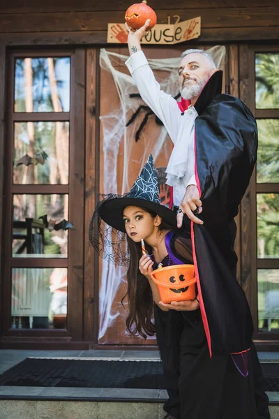 Man in halloween vampire costume holding carved pumpkin in raised hand near daughter in witch hat — Stock Photo