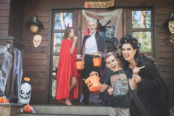 Мати і син посміхаються біля розмитої сім'ї в костюмах Хеллоуїна на прикрашеному ганку — стокове фото