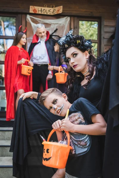 Мама и сын в жутких костюмах на Хэллоуин смотрят в камеру рядом с семьей на размытом крыльце — стоковое фото