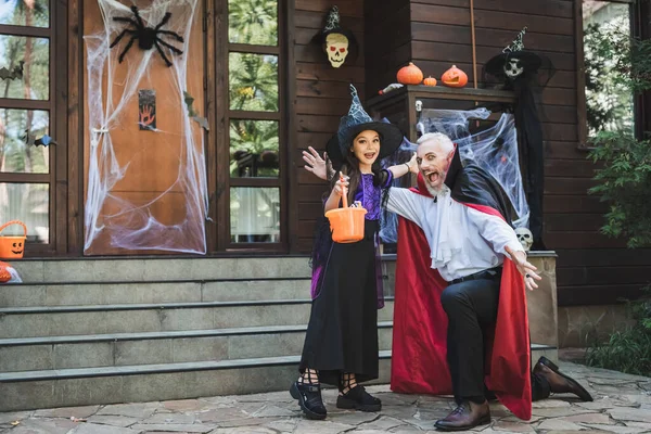 Homem animado em traje de vampiro e menina com balde de doces perto do alpendre com decoração de Halloween — Fotografia de Stock