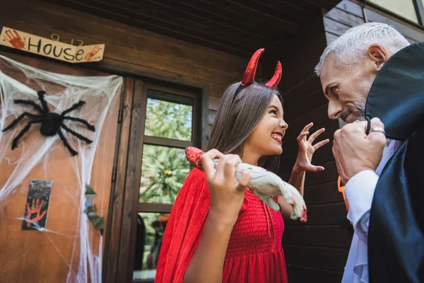 Cheerful girl in devil halloween costume scaring dad with toy hand near cottage with decoration — Stock Photo