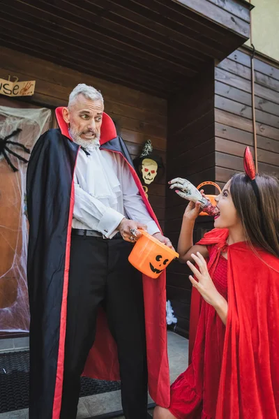 Spooky girl in halloween costume scaring dad with toy hand near house — Stock Photo