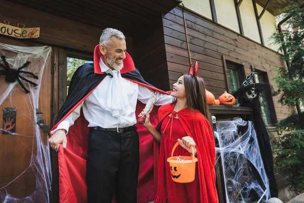 Hombre emocionado cerca de hija feliz en traje de diablo en el porche de la casa - foto de stock