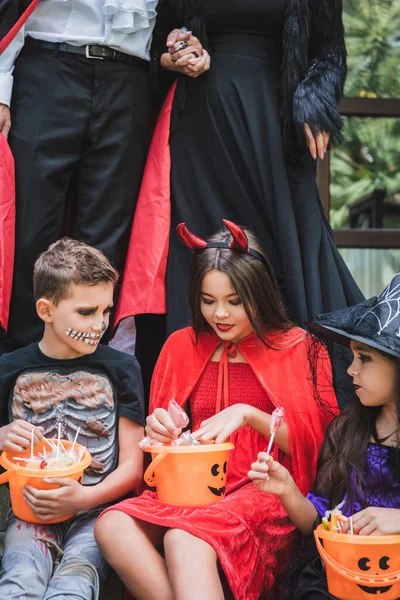 Niños en trajes de Halloween miedo sentado con cubos de piruletas cerca de los padres tomados de la mano en el fondo - foto de stock