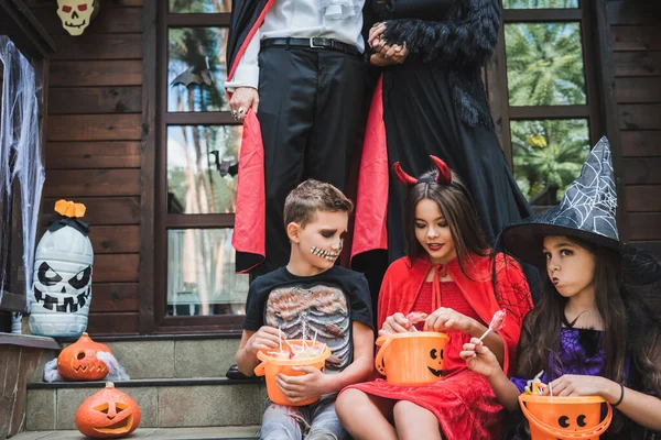 Kids in halloween costumes sitting on cottage stairs with buckets of sweets near parents — Stock Photo
