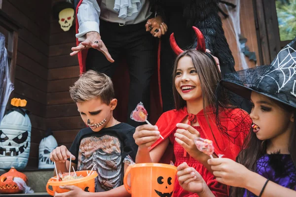 Niños felices en trajes de Halloween sosteniendo cubos con piruletas cerca de los padres — Stock Photo