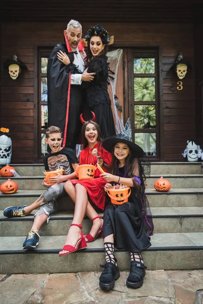 Родители в костюмах на Хэллоуин смотрят на детей, сидящих на коттеджной лестнице с ведрами сладостей — стоковое фото