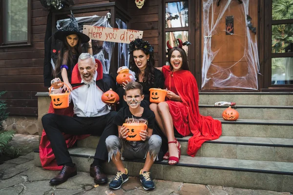 Parents et enfants en costumes d'Halloween effrayants grimacant tout en étant assis sur des escaliers de chalet — Photo de stock