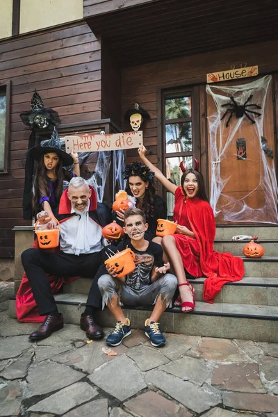 Niños espeluznantes sosteniendo la tarjeta con aquí usted muere letras cerca de los padres en trajes de Halloween - foto de stock