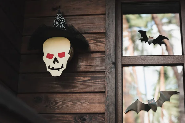Papel cortado cráneo en sombrero de bruja cerca de murciélagos negros en la ventana de la casa de madera - foto de stock