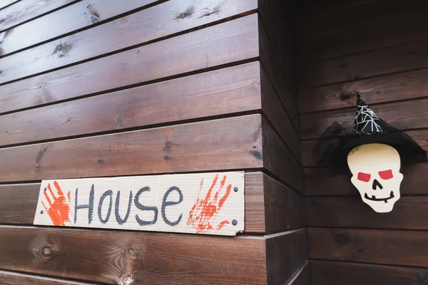 Карточка с надписью дома и кровавыми отпечатками рук рядом с бумагой, разрезанной черепом в шапке ведьмы на деревянном коттедже — стоковое фото