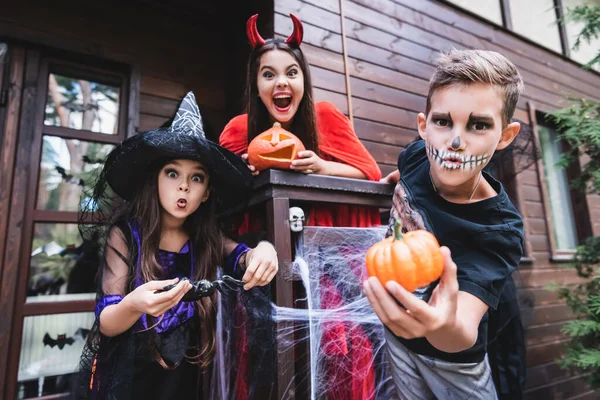 Жуткие детишки в костюмах на Хэллоуин гримасируют на крыльце коттеджа с украшениями — стоковое фото
