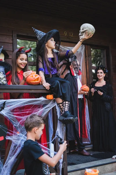 Ragazza in costume di Halloween guardando teschio raccapricciante vicino alla famiglia sul portico con decorazione — Foto stock