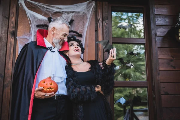 Glücklicher Mann im Vampirkostüm mit geschnitztem Kürbis und umarmender Frau auf Hüttenveranda mit Halloween-Dekoration — Stockfoto