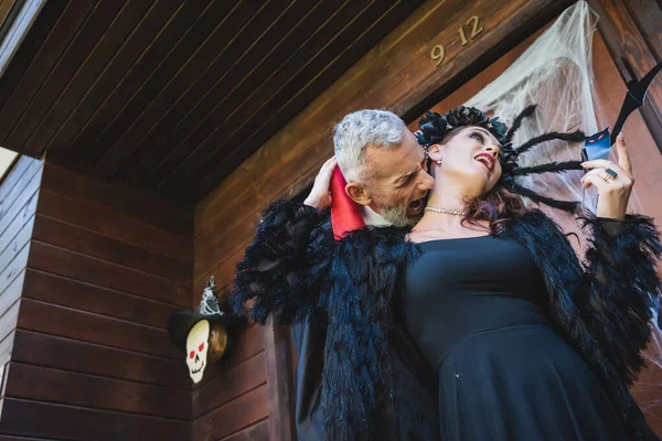 Low angle view of man in halloween vampire costume biting neck of wife on cottage porch — Stock Photo