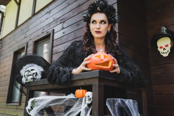 Mujer en traje de vampiro sosteniendo calabaza tallada cerca de valla de porche con decoración de halloween - foto de stock