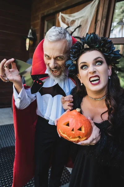 Spooky couple in halloween vampires costumes grimacing with carved pumpkin and paper cut bat — Stock Photo