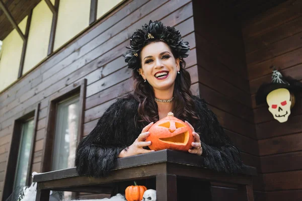Cheerful woman in black halloween dress and wreath standing on wooden balcony with carved pumpkin — Stock Photo
