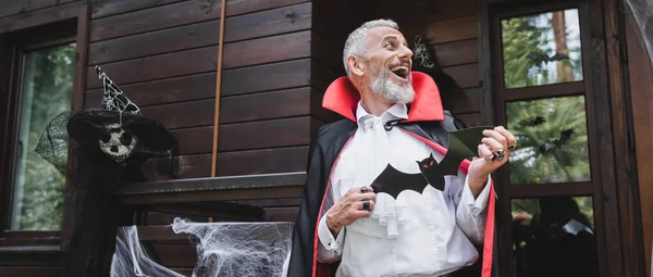 Laughing middle aged man in halloween vampire costume holding paper cut bat on porch of wooden house, banner — Stock Photo