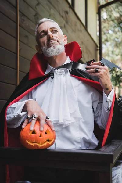 Mature man in vampire halloween costume, with paper cut bat and curved pumpkin — Stock Photo