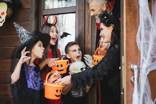 Niños en trajes de Halloween espeluznantes sosteniendo cubos y gruñendo a vecinos sonrientes — Stock Photo