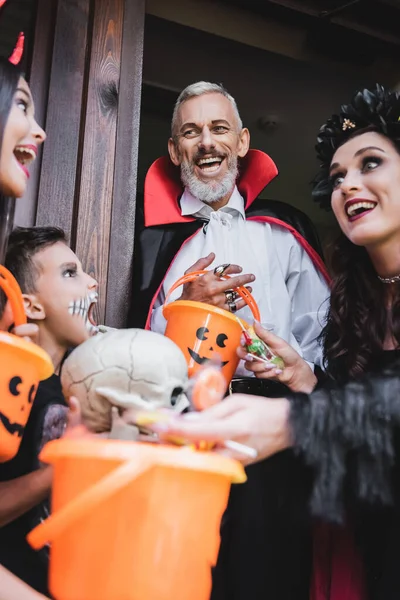 Cheerful couple in halloween costumes smiling at kids near buckets of sweets — Stock Photo