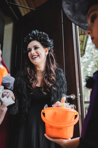 Happy woman in halloween costume near blurred daughter with bucket of sweets — Stock Photo