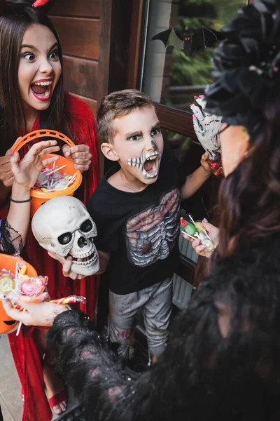 Розмита жінка тримає цукерки поблизу диких дітей у костюмах Хеллоуїна, що ростуть, тримаючи відра — стокове фото