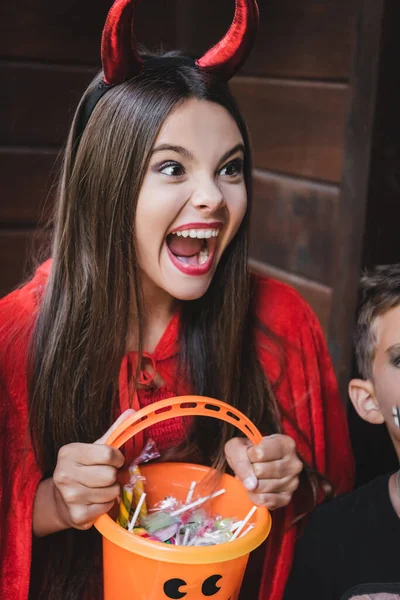 Girl in devil halloween costume holding bucket with sweets and screaming near brother — Stock Photo