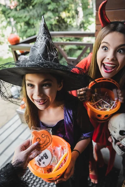 Hermanas sorprendidas en trajes de bruja y diablo cerca de la madre con galleta de halloween - foto de stock