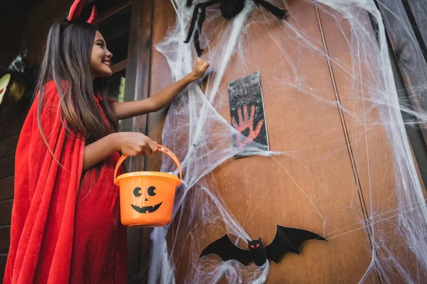Low angle view of cheerful girl in devil costume knocking door with halloween decoration — Stock Photo