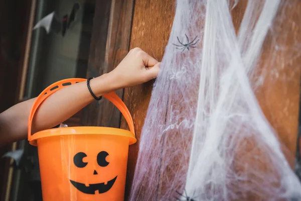 Vista recortada de niña sosteniendo cubo con cara espeluznante pintada mientras llama a la puerta cerca de la red de araña decorativa - foto de stock