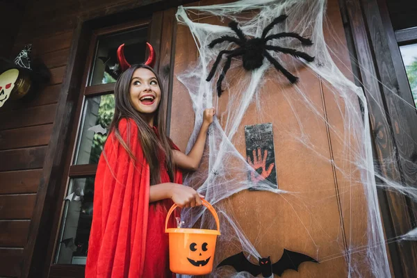 Menina animado em traje do diabo batendo em portas com decoração halloween — Fotografia de Stock