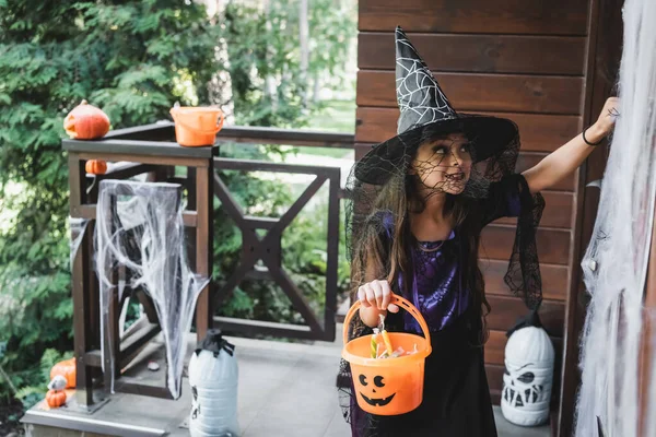 Child in witch halloween costume holding bucket with sweets while knocking at door on decorated porch — Stock Photo