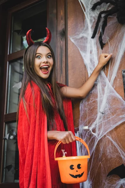 Aufgeregtes Mädchen im Teufel-Halloween-Kostüm hält Eimer und klopft mit Spinnennetz an Tür — Stockfoto