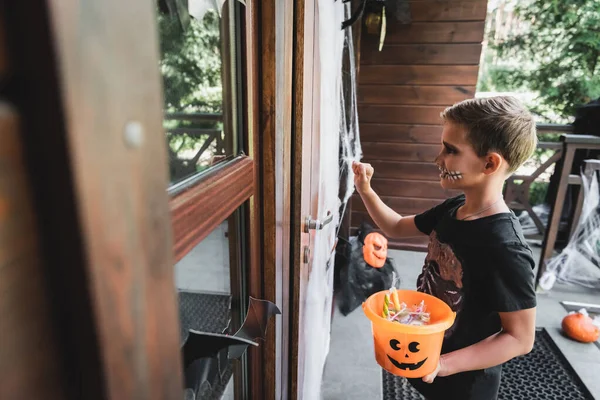 Хлопчик у скелетному костюмі тримає відро Хеллоуїна, стукаючи у двері котеджу — стокове фото