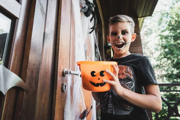 Arrabbiato ragazzo in costume scheletro tenendo secchio di Halloween e ringhiando vicino porta cottage — Foto stock