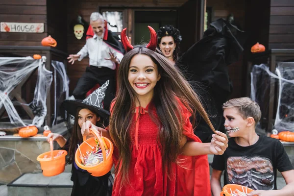 Glückliches Mädchen im Teufel-Halloween-Kostüm mit einem Eimer voller Süßigkeiten, während es mit Freunden aus dem Nachbarhaus rennt — Stockfoto