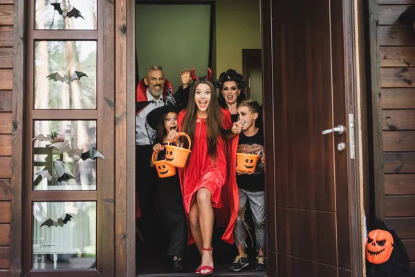 Amigos emocionados en disfraces de Halloween gritando mientras se agota casa vecina con cubos de dulces - foto de stock