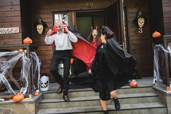 Scared couple in halloween costumes running out cottage from spooky and shouting girl — Stock Photo